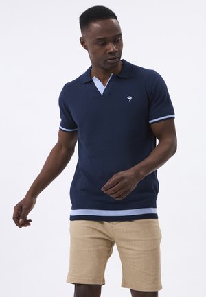 Mann trägt eine marineblaue kurzärmelige Poloshirt mit hellblauer Paspel und beigefarbenen Shorts, schaut nach unten und leicht zur Seite.