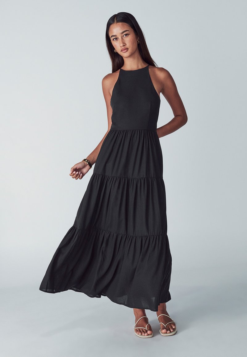 Mladá žena v černých maxi šatech bez rukávů s vrstvením a s sandály na zavazování, stojící proti jednoduchému světle šedému pozadí.