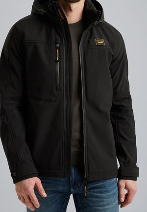 Homme portant une veste noire zippée avec une poche poitrine zippée et des logos PME Legend, associée à un jean bleu et un pull sombre en dessous.