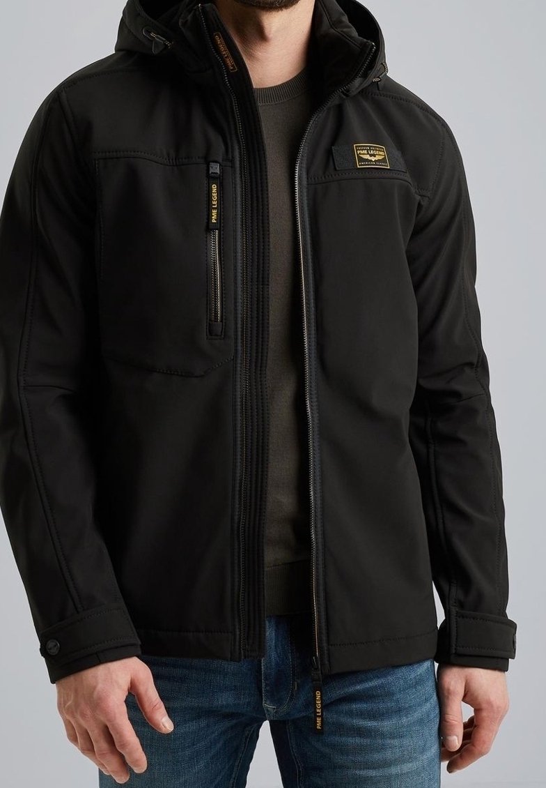Homme portant une veste noire zippée avec une poche poitrine zippée et des logos PME Legend, associée à un jean bleu et un pull sombre en dessous.