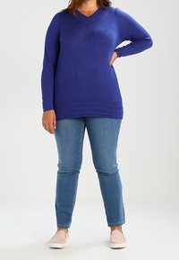 Personne portant un pull bleu à manches longues, un jean bleu et des chaussures légères roses, debout avec une main sur la hanche.