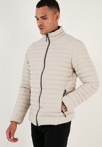 Lyse beige pufjakke med lodret syning, høj krave og lynlåslommer. Har elastiske manchetter og et strømlinet design.