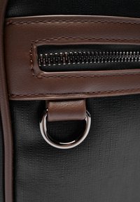 Tissu noir texturé avec des accents en cuir marron, une poche zippée et un anneau en D métallique. Des coutures marron mettent en valeur les détails de conception.
