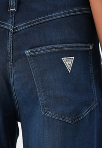 Tmavě modré džínové kalhoty mají přišitou zadní kapsu s trojúhelníkovou visačkou "GUESS". Látka má hladkou strukturu.