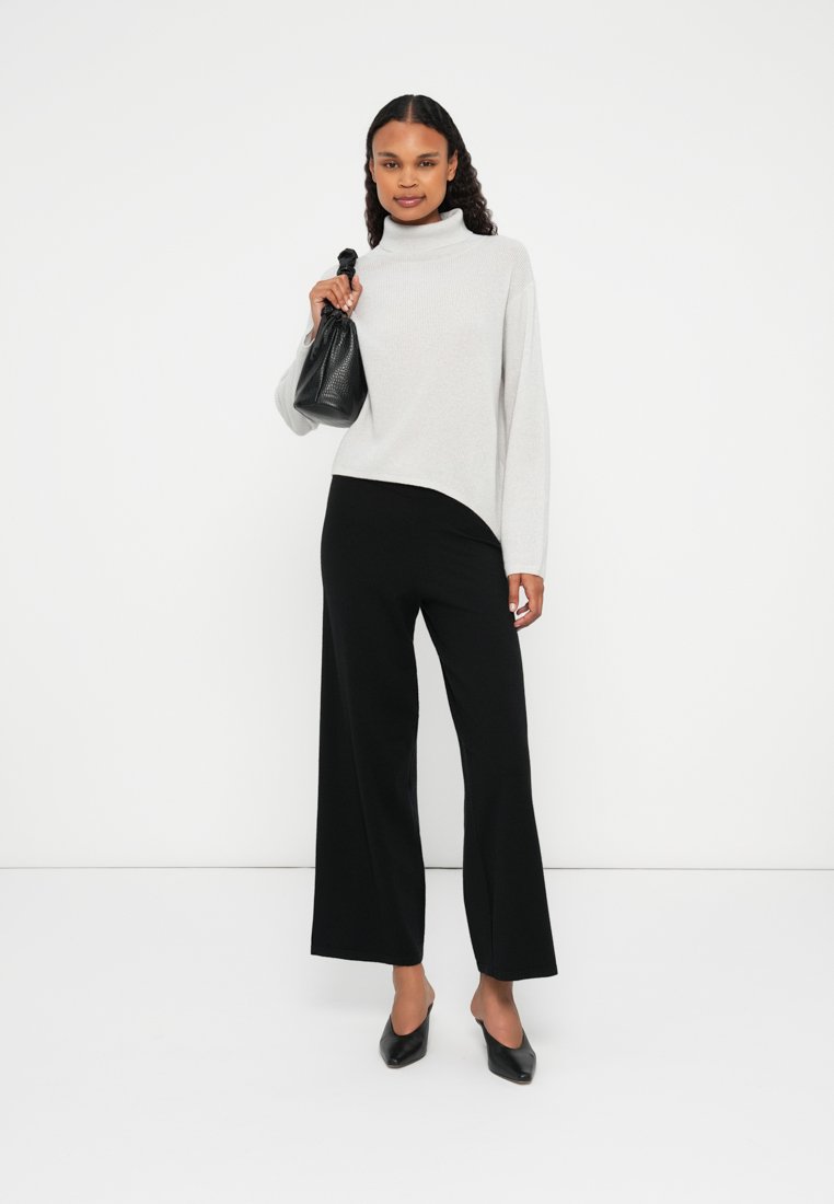 Un pull à col roulé gris clair avec une texture côtelée, associé à un pantalon noir à jambes larges. La mannequin tient un petit sac noir et porte des chaussures pointues noires.