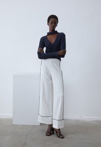 Top de manga larga de canalé en navy con un corte triangular, combinado con pantalones anchos blancos con ribete negro y un dobladillo limpio. Zapatos de tacón negro.