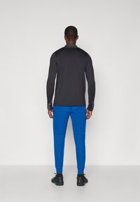 Blusa preta de manga longa e calças de treino azuis. A blusa possui uma textura suave; as calças têm punhos elásticos e um corte ajustado. Sapatos pretos.