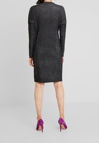 Abito nero in maglia con una texture luccicante, maniche lunghe e taglio al ginocchio, abbinato a scarpe con tacco alto viola.