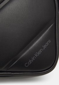 Borsa in pelle sintetica nera con texture e design angolare. Presenta il marchio "Calvin Klein Jeans" in un colore chiaro sul lato.