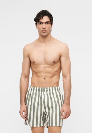 Άνδρας με σκούρα σγουρά μαλλιά, που φοράει γκρι και λευκά κάθετα ριγέ μαγιό, στέκεται μπροστά σε απλό φόντο.