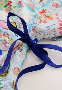 Barevná látka s pobřežním motivem obsahujícím mušle a hvězdice, uvázaná tmavě modrou stuhou na krku. Měkký, texturovaný materiál.