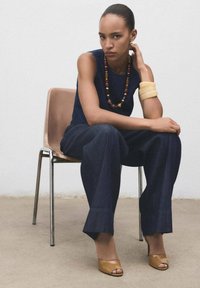 Haut sans manches bleu marine, large pantalon en denim foncé, chaussures à talons ouvertes marron, collier multirangs avec des perles en bois, et bracelets épais au bras.