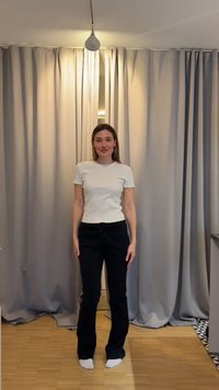 Žena stoji bosa na drvenom podu, nosi bijelu majicu kratkih rukava i crne hlače, ispred zatvorenih sivih zavjesa.