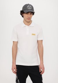 Bela polo majica s ovratnikom in tremi gumbi, na levem prsnem delu ima majhen zlat logotip. Črna kapa z detajlom logotipa.