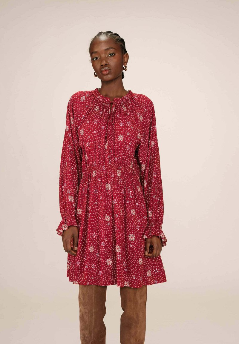 Robe rouge à motifs, avec des manches longues, un col froncé et des motifs floraux ; présente une coupe décontractée et des poignets volantés, portée avec des bottes montantes.