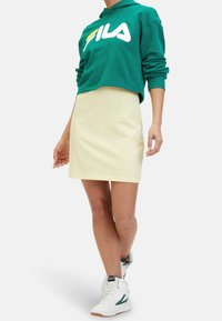 Persona che indossa un hoodie verde corto con un logo bianco, una gonna crema e sneaker alte bianche, in piedi con una mano sui fianchi.