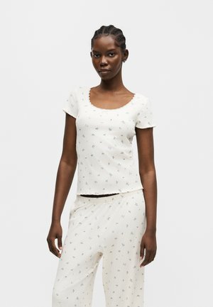 Nő fehér rövid ujjú felsőt és hozzáillő nadrágot visel, apró kék virágmintával, áll egy egyszínű világos háttér előtt.