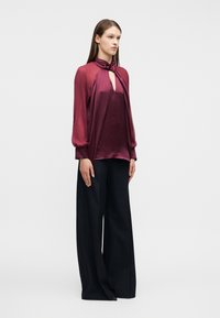 Γυναίκα που στέκεται, φοράει μπλούζα σατέν μπορντό με μακριά μανίκια, δεμένη στη λαιμόκοψη, και φαρδιά μαύρη παντελόνα, πάνω σε απλό φόντο.