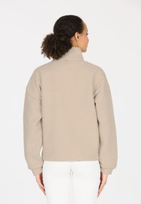 Beige Fleecejacke mit hohem Kragen, strukturierten Ärmeln und weicher Textur. Verfügt über ein kleines Markenetikett auf der Rückseite.