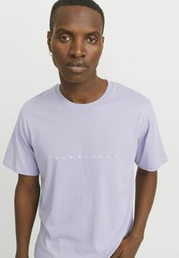 Levandų spalvos trumpomis rankovėmis marškinėliai, pagaminti iš minkštos medvilnės; priekį puošia balta spalva užrašyta prekės ženklą "JACK & JONES".
