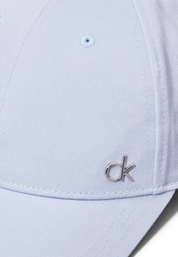 Lyseblå caps laget av teksturert stoff, med et sølvfarget "ck"-logo og strukturert skjerm, med sømdetaljer og ventilasjonshull.