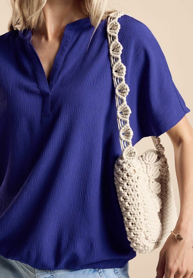 Femme portant un chemisier bleu roi texturé et un jean bleu clair, portant un sac à épaule en crochet couleur crème avec des motifs floraux.