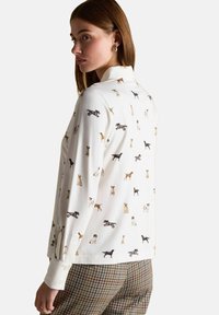 Žena v bielom tričku s dlhým rukávom a farebnými ilustráciami psov, skombinovaná s hnedými károvanými nohavicami, zobrazená zozadu z boku.