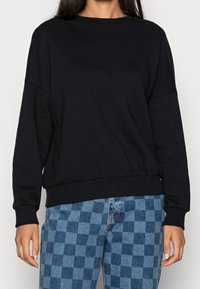 Felpa nera in cotone con collo rotondo, maniche lunghe e orlo a costine. Abbinata a jeans con motivo a quadri blu.