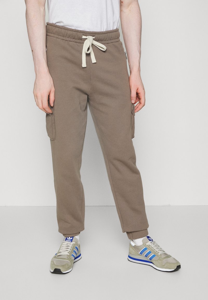 Homme portant un pantalon de jogging cargo marron clair avec taille à cordon et des baskets beiges à rayures bleues, debout devant un fond clair uni.