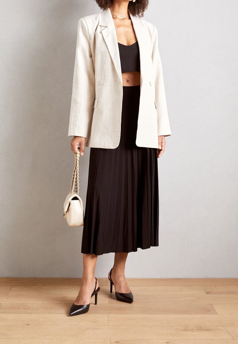 Femme portant un blazer beige, un haut court noir, une jupe midi noire plissée, des talons noirs, et tenant un sac à main beige avec une bandoulière en chaîne dorée.