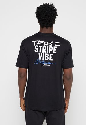 Hombre con pelo corto y rizado que lleva una camiseta negra con texto blanco y azul "TRIPLE STRIPE VIBE" en la espalda, de pie frente a un fondo liso.
