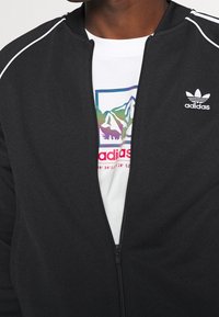 Chaqueta negra con cremallera y ribetes blancos, con un logo en el pecho y superpuesta a una camiseta blanca que muestra un gráfico colorido de montañas.
