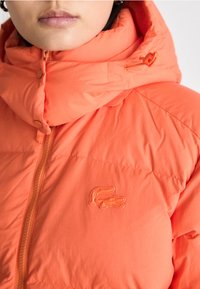 Narančasta puffer jakna s visokim ovratnikom, koja ima glatku teksturu, patentni zatvarač sprijeda i izvezeni logotip na prsima.