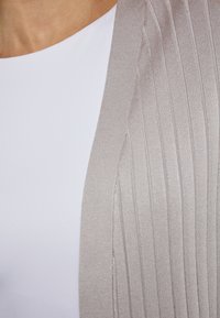 Licht beige geribde cardi drapeert over een witte top, met een zachte textuur en een open ontwerp zonder knopen of sluitingen.
