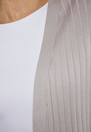 Licht beige geribde cardi drapeert over een witte top, met een zachte textuur en een open ontwerp zonder knopen of sluitingen.