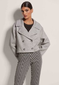 Gekürzte, doppelt geknöpfte graue Jacke mit großen Revers. Kombiniert mit gemusterten schwarz-weißen Hosen, die geometrische Formen aufweisen.