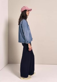 Kvinde i pink kasket, denimjakke, sorte vidde bukser og beige sneakers står sidelæns mod en ensfarvet beige væg.