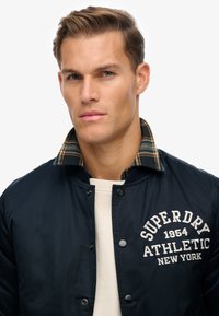 Chaqueta de piloto marina con cierre a presión, que presenta un cuello de cuadros y un logo bordado en blanco. Tela suave y duradera con un ajuste entallado.