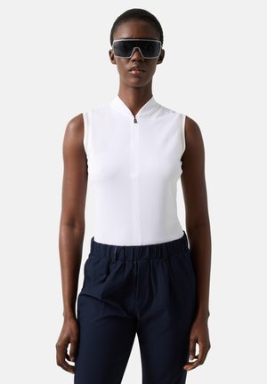 Kobieta w białej, bezrękawnikowej bluzce na zamek z przodu, ciemnych spodniach z wysokim stanem i dużych czarnych okularach przeciwsłonecznych, stojąca na jednolitym tle.