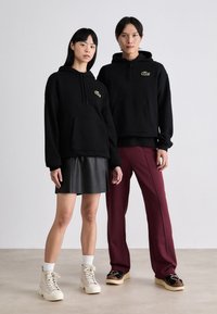 Sudaderas negras con bolsillos canguro, con un logo verde, combinadas con una falda de cuero y pantalones color burdeos. Calzado blanco y beige.