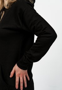 Černá mikina s dlouhým rukávem a přední kapsou, s volným střihem a hladkou bavlněnou texturou. Viditelné švy podél spojů.