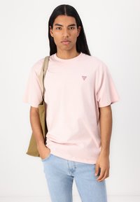 Camiseta de algodón de color rosa claro con cuello redondo, manga corta y un pequeño logo triangular rojo en el lado izquierdo del pecho. Combinada con unos vaqueros de color azul claro.