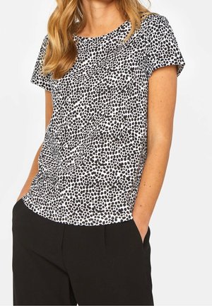 Femme portant un t-shirt blanc à manches courtes avec un motif de points noirs irréguliers et un pantalon noir, les mains dans les poches, devant un fond blanc.