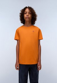 T-shirt en coton orange avec un col rond et des manches courtes, arborant un petit écusson du drapeau norvégien. Associée à un jean foncé.