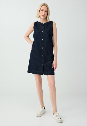 Donna bionda in piedi con una mano sui fianchi che indossa un vestito in denim scuro senza maniche con bottoni sul davanti e sneakers bianche su sfondo uniforme.