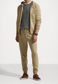 Mężczyzna ubrany w beżową bluzę z kapturem i spodnie dresowe, pod spodem szara koszulka, brązowe trampki oraz zegarek z brązowym skórzanym paskiem, stojący na białej podłodze.