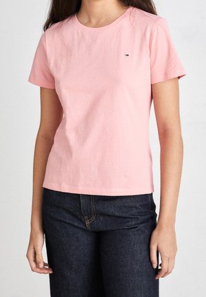 Kobieta w gładkim, jasnoróżowym, krótkim rękawie oraz ciemnoniebieskich dżinsach, stojąca na jednolitym tle.