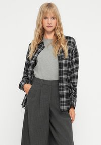 Chemise à carreaux noir et blanc sur un t-shirt gris, associée à un pantalon large taille haute gris foncé. Tissu texturé et coupe décontractée.