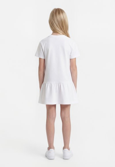 Νεαρή κοπέλα με μακριά ξανθά μαλλιά στέκεται με την πλάτη προς τα μέσα φορώντας ένα λευκό φόρεμα με κοντά μανίκια και βολάν στο κάτω μέρος, καθώς και λευκά αθλητικά παπούτσια.