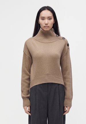 Femme portant un pull à col roulé en maille beige avec des boutons sur l'épaule et un pantalon large foncé, debout devant un fond uni.